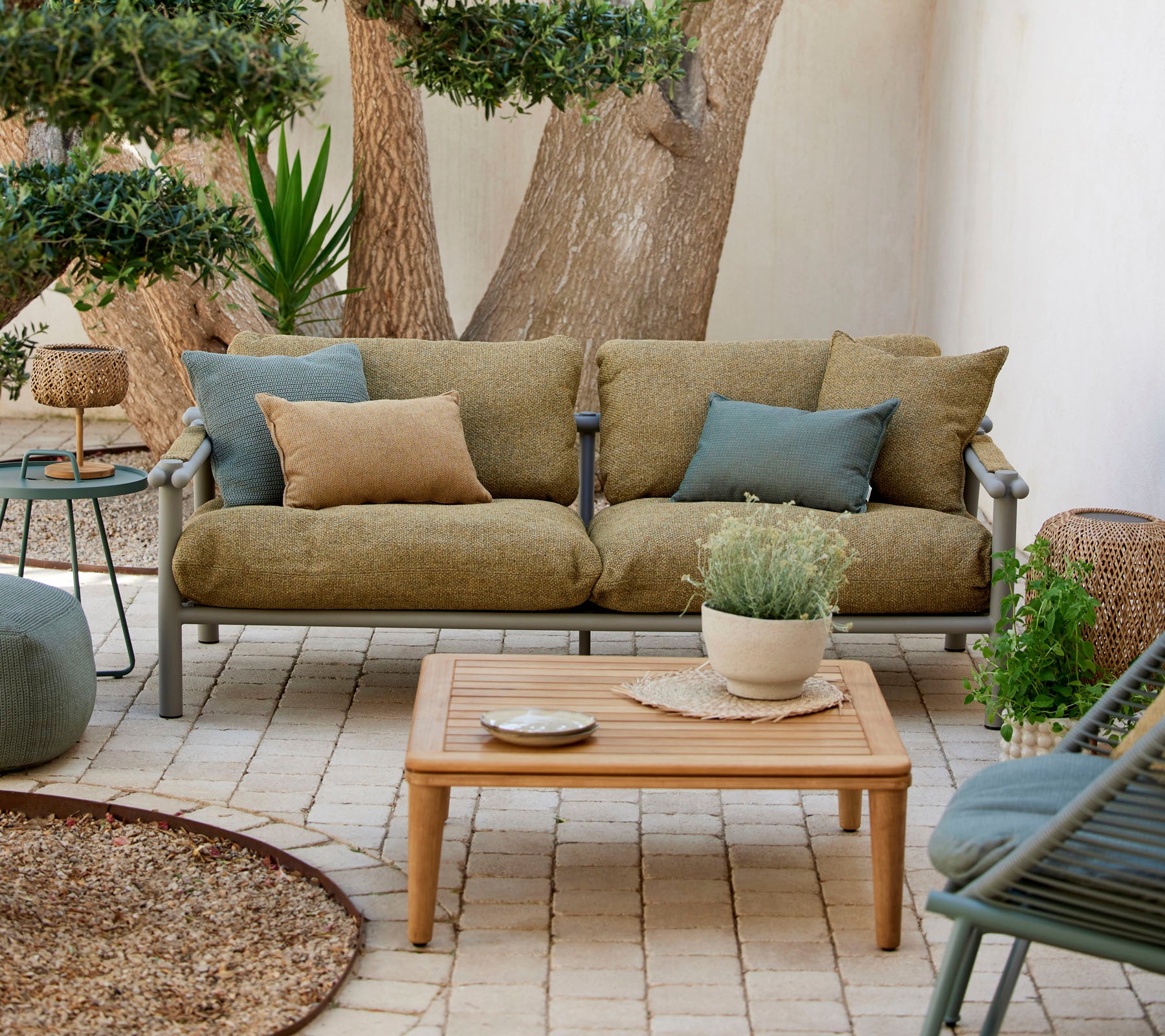Zona de estar cómoda con un sofá verde con cojines, una mesa de café de madera y plantas decorativas en un entorno sereno.