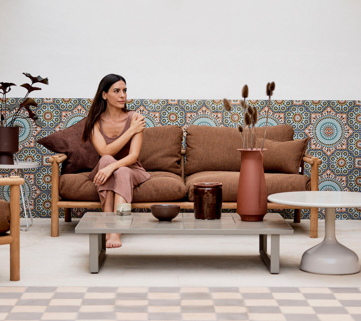 Una mujer sentada en un sofá con cojines decorativos, rodeada de jarrones y plantas, contra un fondo estampado.