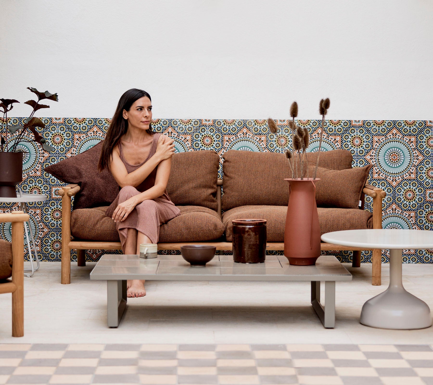 Una mujer sentada en un sofá con cojines decorativos, rodeada de jarrones y plantas, contra un fondo estampado.