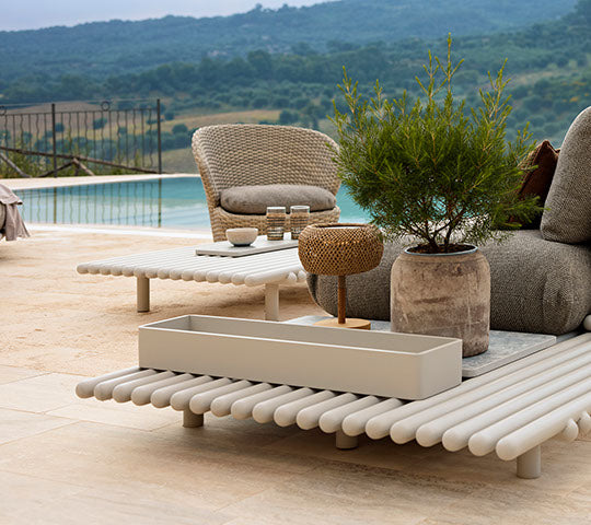 Mobiliario exterior elegante con varillas de aluminio, una maceta redonda y elementos decorativos, contra un paisaje sereno.