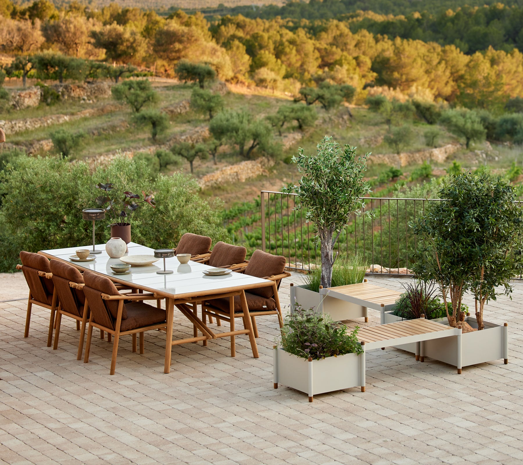 Conjunto de comedor de teca estiloso con sillas y un banco para plantas, rodeado de verdor y un paisaje pintoresco.