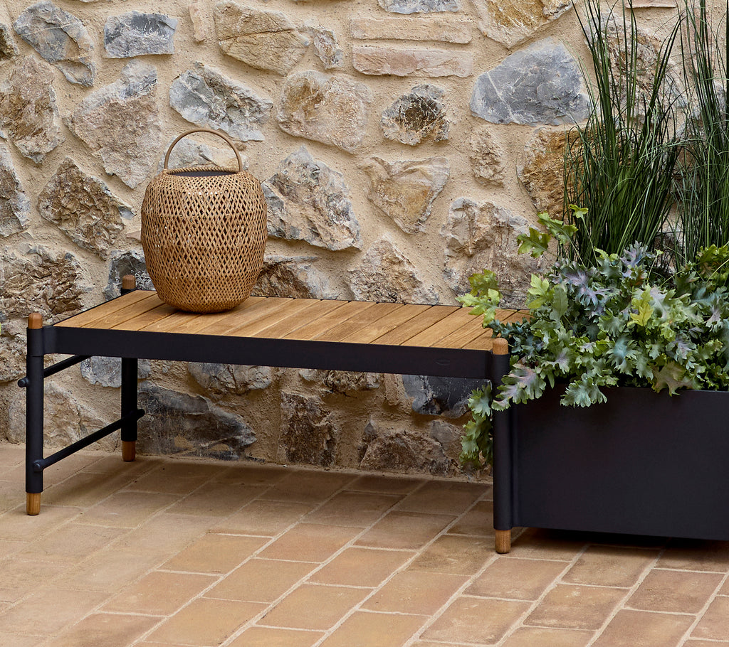 Un banco elegante con una parte superior de madera, acompañado de una cesta trenzada y una maceta decorativa llena de vegetación contra una pared de piedra.