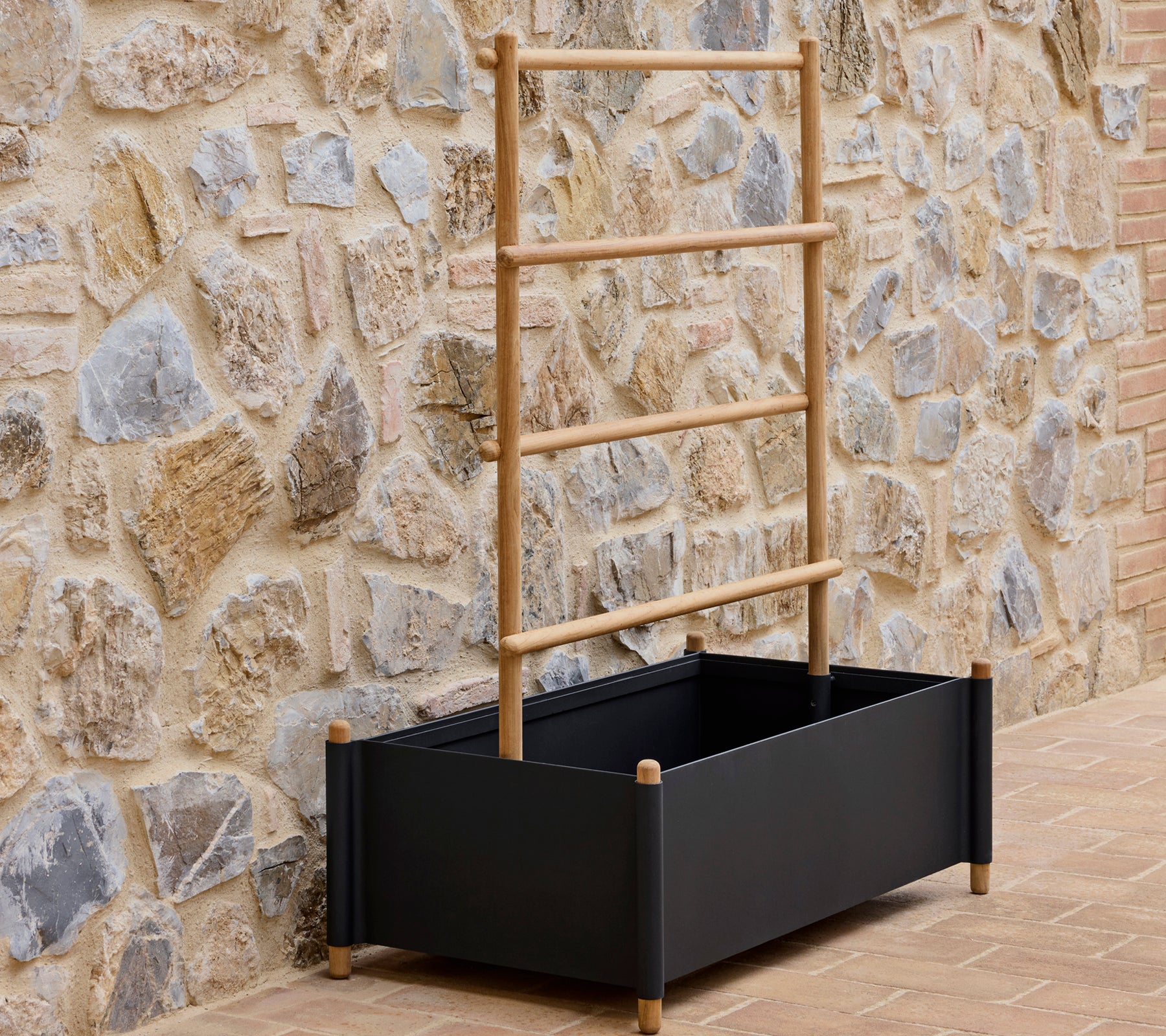 Maceta de aluminio negro con varillas de soporte de madera, colocada contra una pared de piedra texturizada.