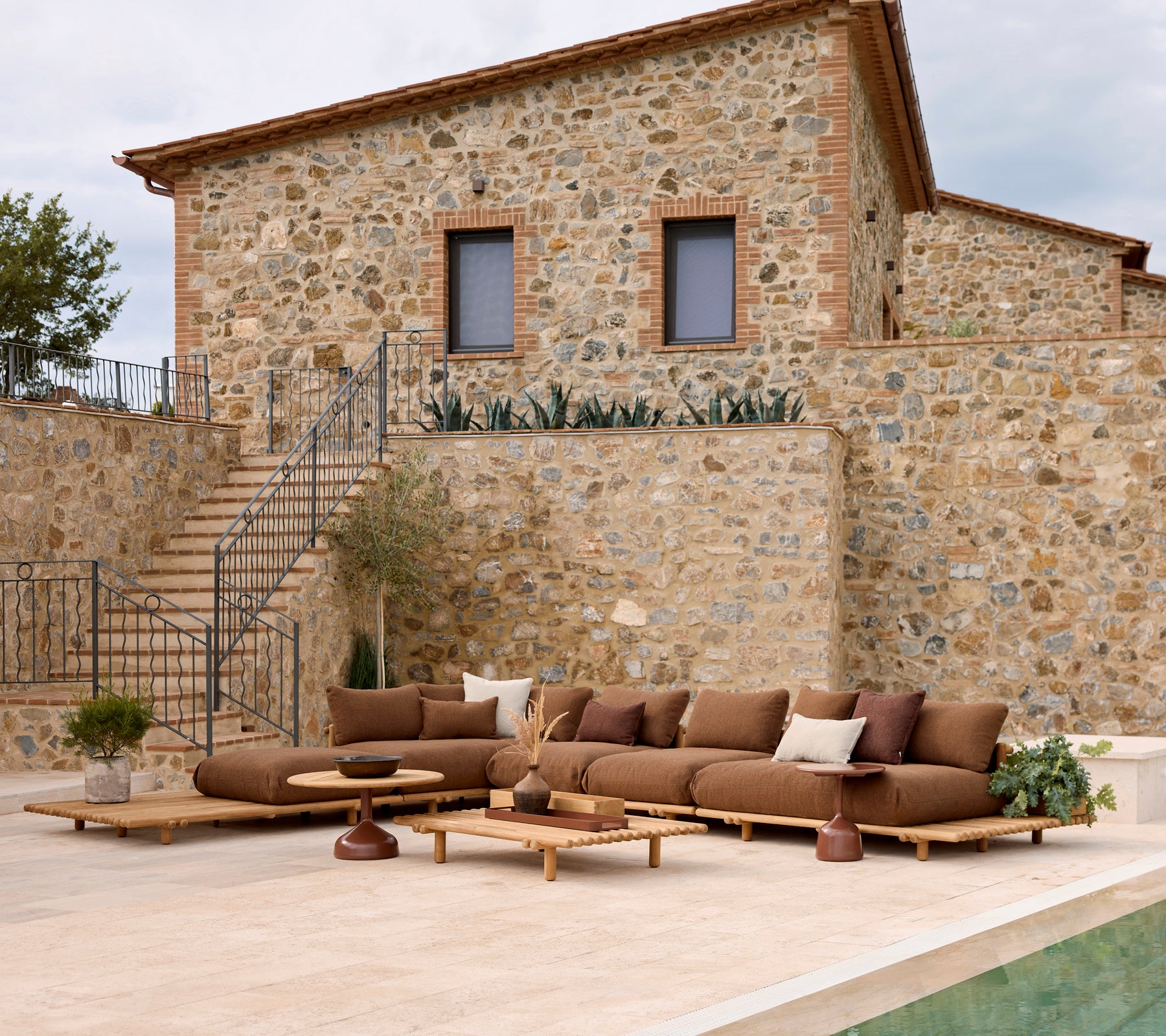 Sofá lounge con cojines suaves al lado de una mesa de café elegante, frente a una pared de piedra. Las macetas añaden un toque natural.
