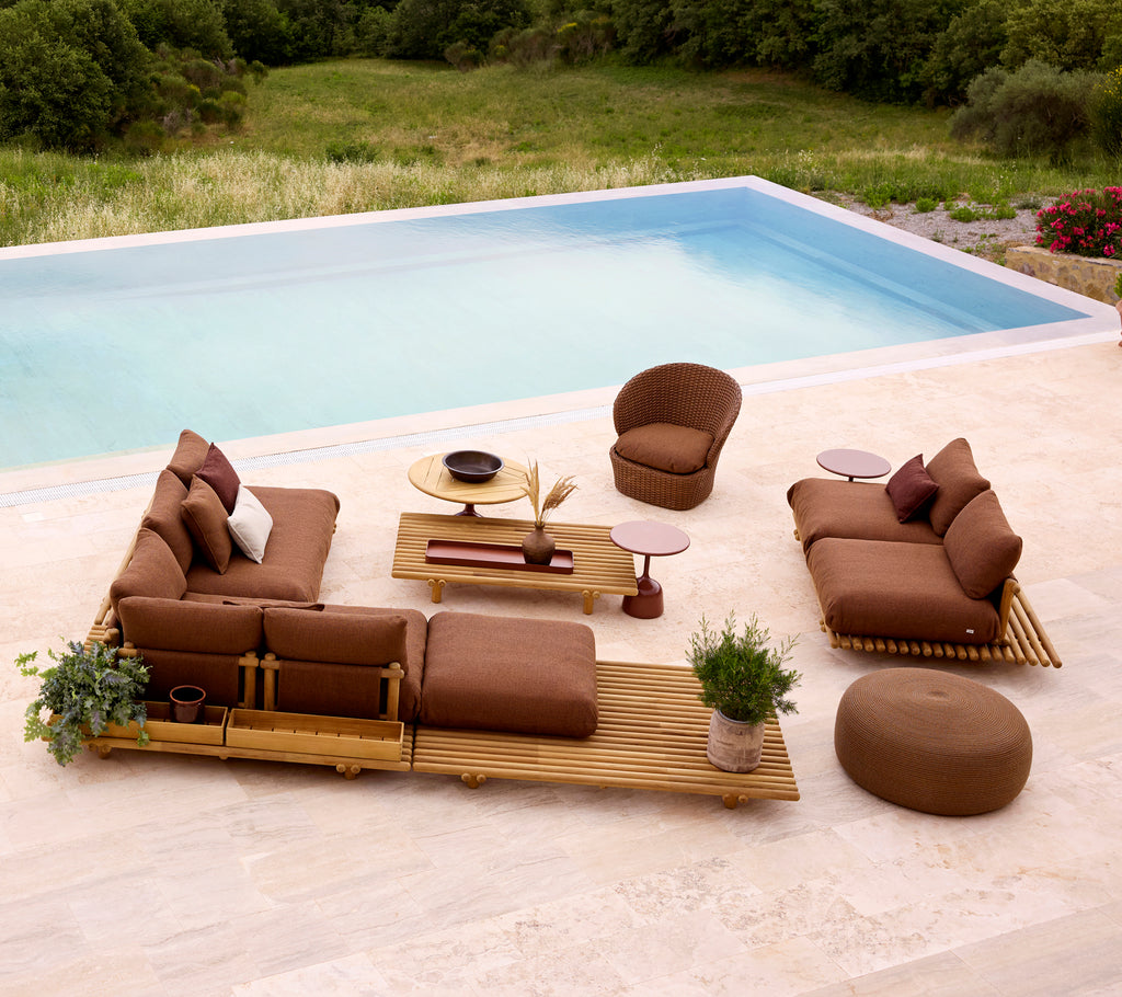 Configuración de lounge elegante con muebles de teca, cojines suaves y un reposapiés redondo, complementada con vegetación y una mesa.
