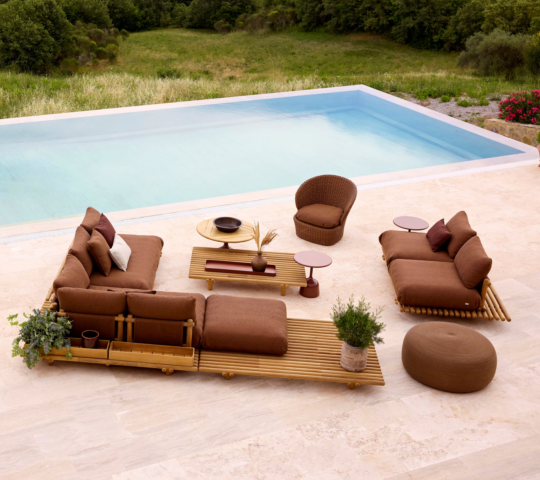 Configuración de lounge elegante con muebles de teca, cojines suaves y un reposapiés redondo, complementada con vegetación y una mesa.