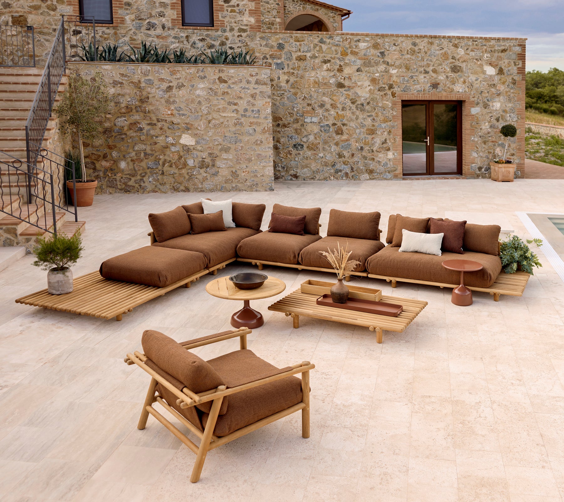 Una zona de salón elegante con un sofá seccional con cojines suaves, una mesa de café y una silla de lounge de teca en un fondo de piedra.