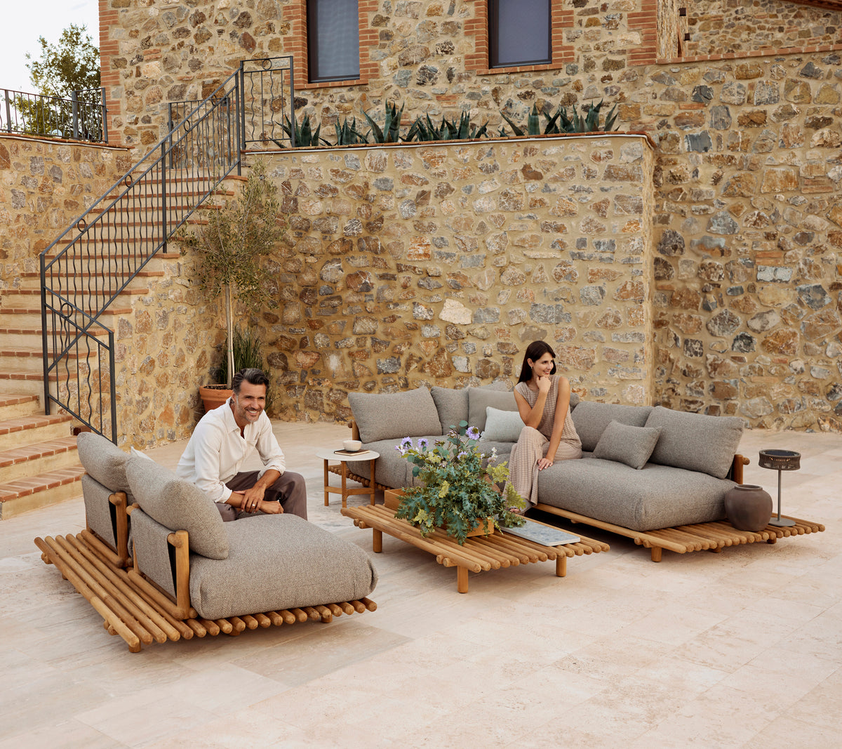 Un área de salón elegante con muebles de teca y cómodos cojines grises, acentuada por vegetación y una mesa de café moderna.