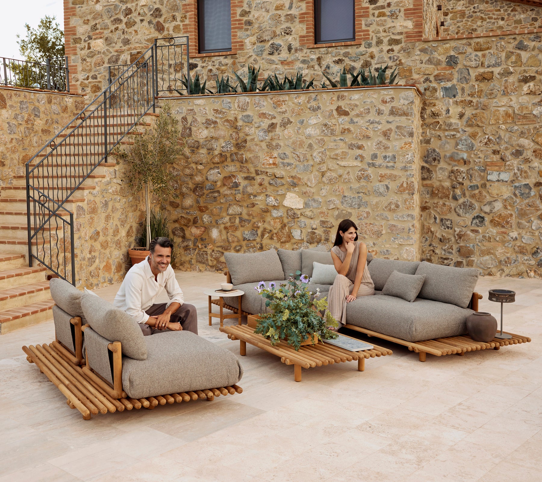 Un área de salón elegante con muebles de teca y cómodos cojines grises, acentuada por vegetación y una mesa de café moderna.