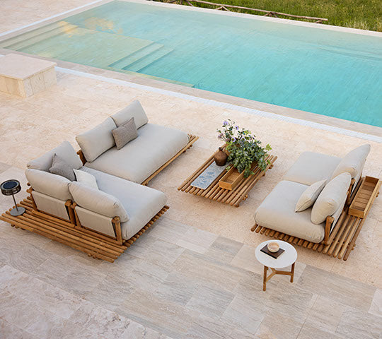Mobiliario de teca elegante con cojines Natte, complementado con una mesa de café a juego y una planta en maceta, situada al lado de una piscina tranquila.
