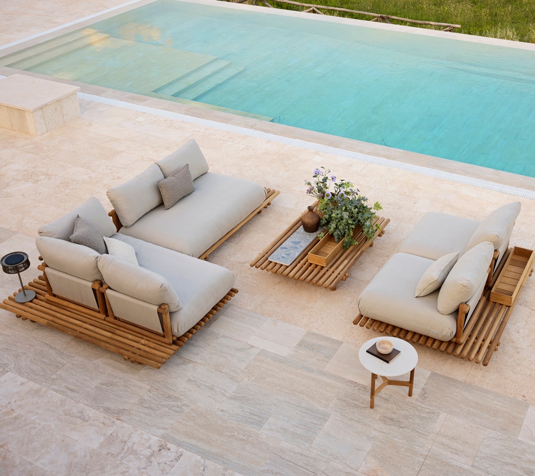 Área de descanso exterior moderna con cojines neutros, mesas de plataforma de madera y una pequeña planta en maceta junto a una piscina.