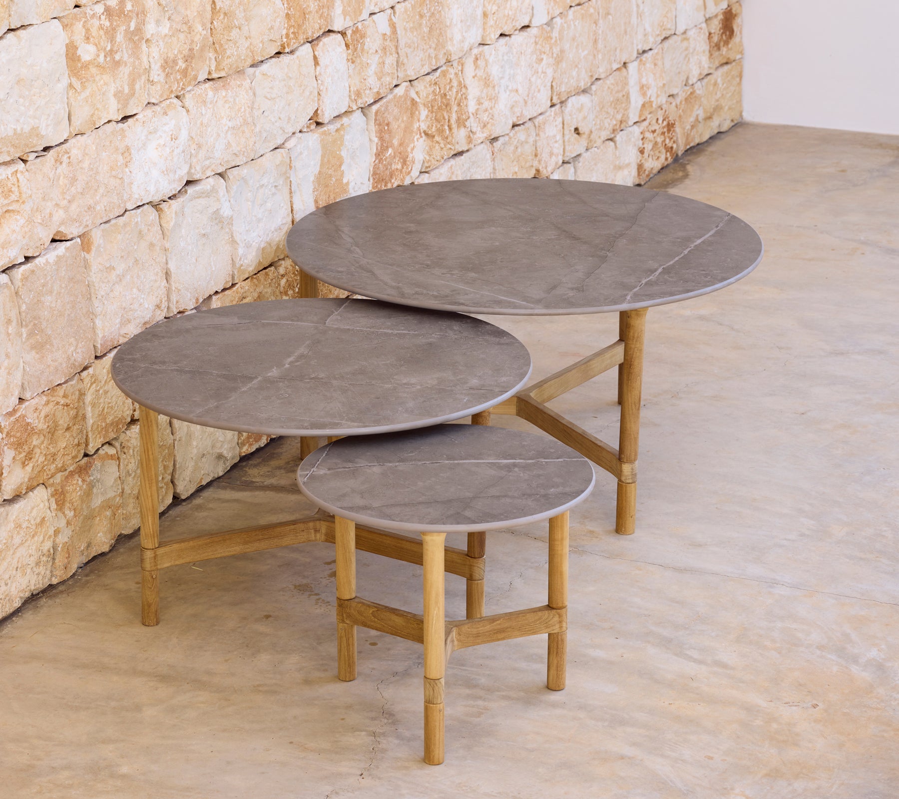 Tres mesas de café grises modernas con bases de madera clara contra una pared de piedra.