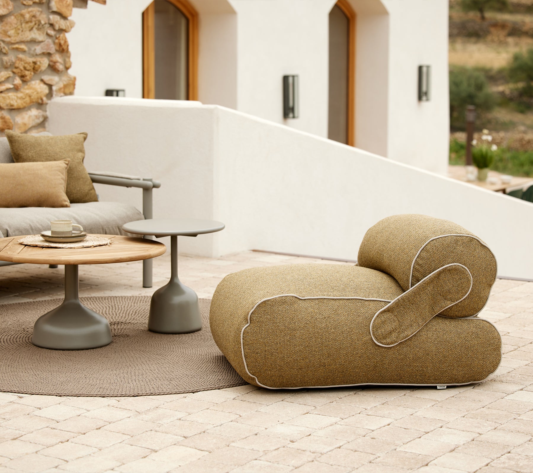 Sillón elegante con una forma única, acompañado de una mesa redonda y cojines decorativos, situado en un entorno tranquilo.