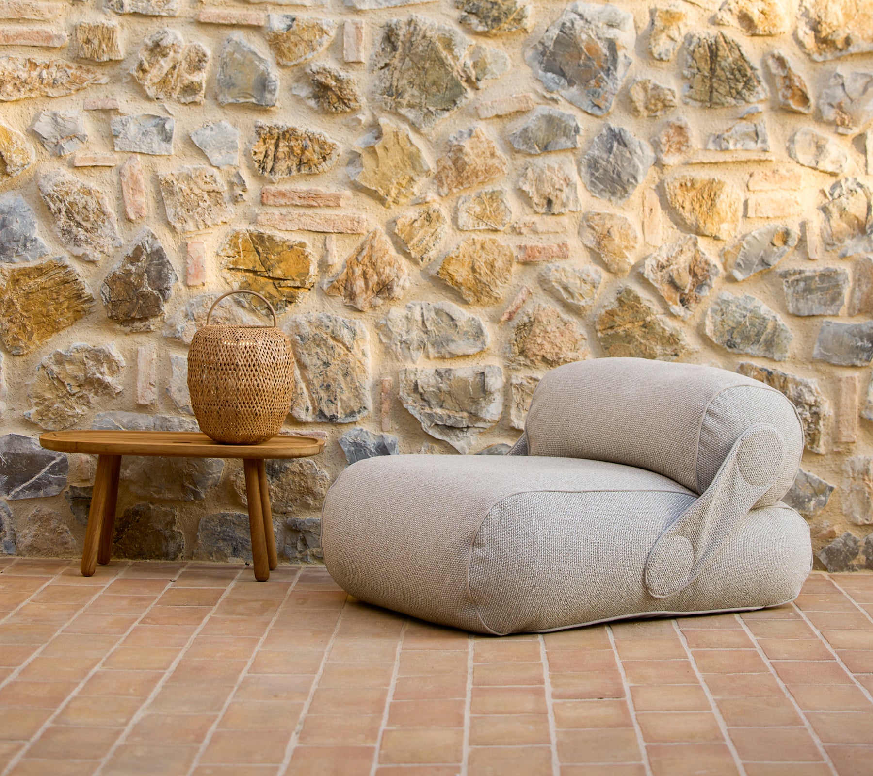 Una silla lounge elegante al lado de una mesa de café de madera y un jarrón decorativo, situada frente a una pared de piedra rústica.