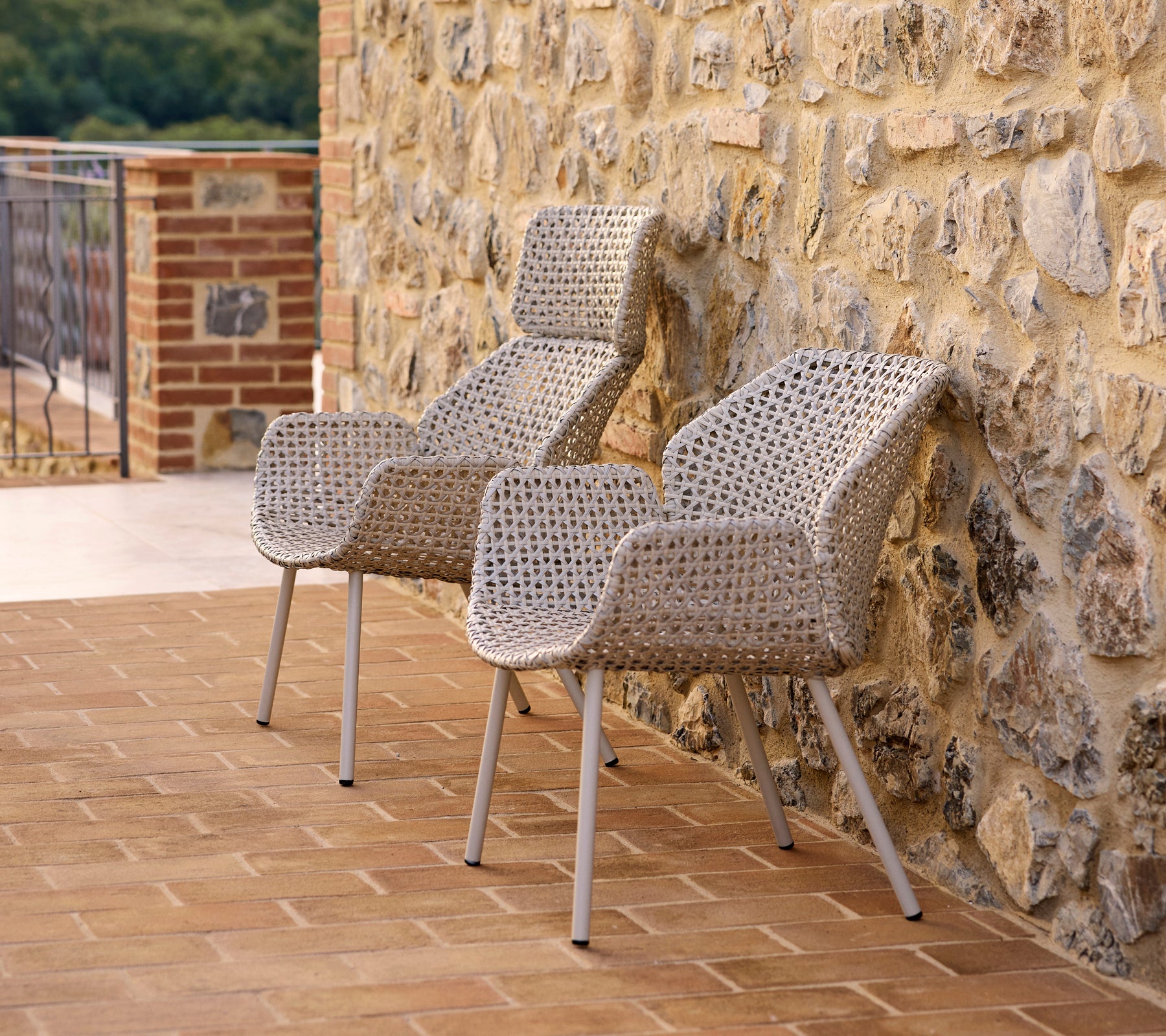 Dos elegantes sillones de respaldo alto Vibe hechos de material tejido, ubicados contra una pared de piedra, perfectos para relajarse.