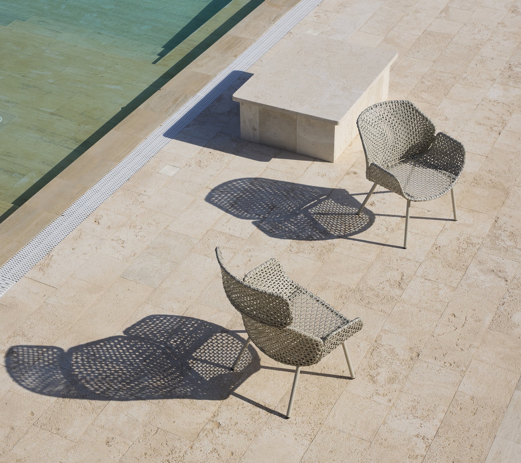 Dos elegantes sillones con diseños únicos que proyectan sombras, situados cerca de una superficie de agua tranquila y una mesa de piedra.