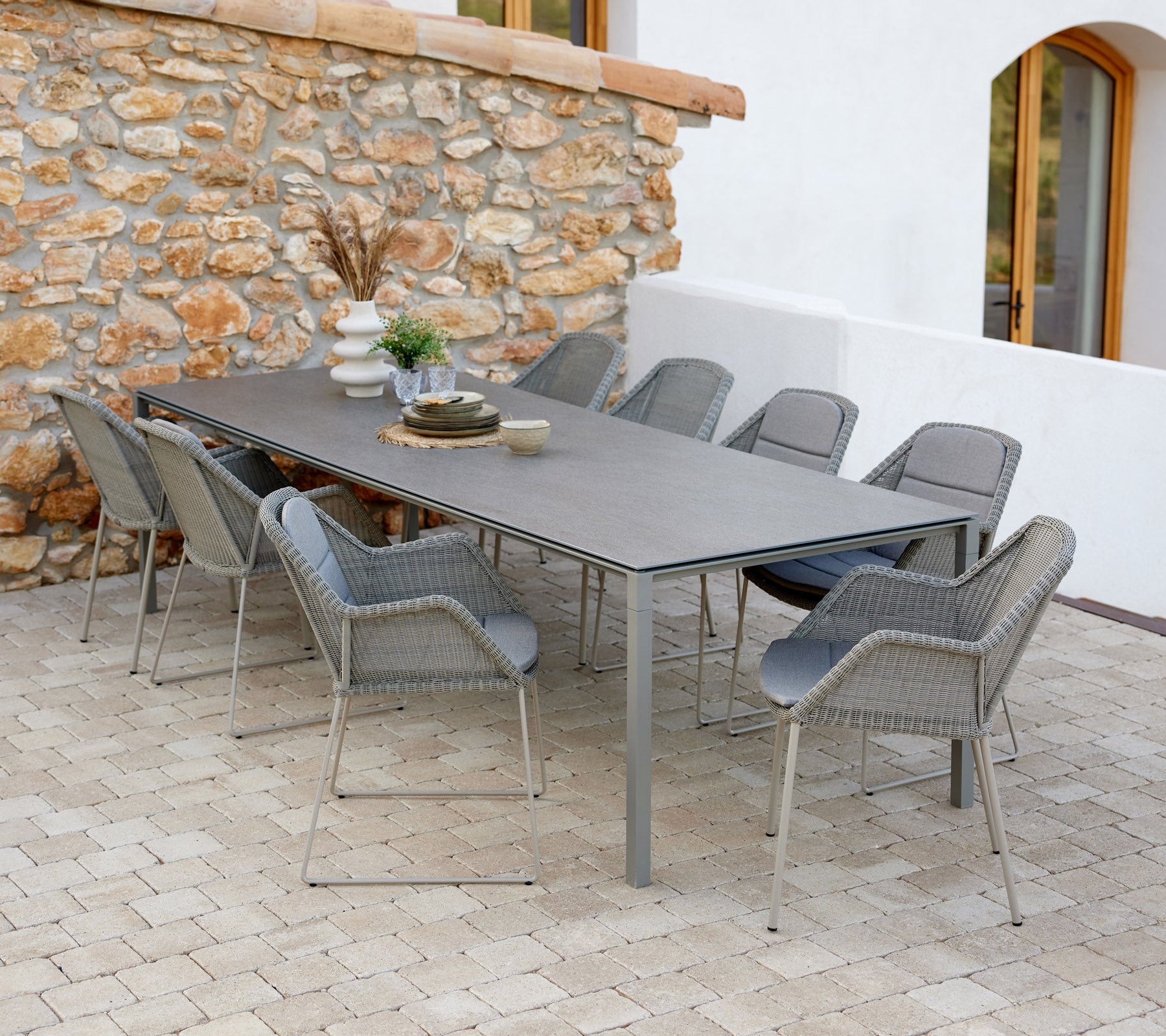 Mesa de comedor moderna con sillas taupe, decorada con elementos decorativos y un toque de verdor contra un fondo de pared de piedra.