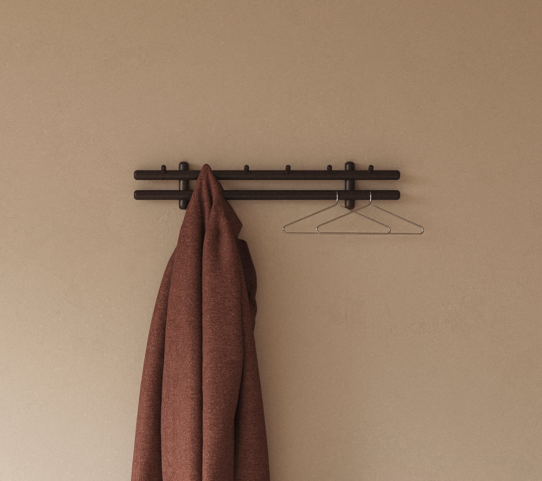 Un abrigo marrón cuelga de un estante de pared minimalista con ganchos, complementado por una simple percha de alambre contra un fondo neutro.
