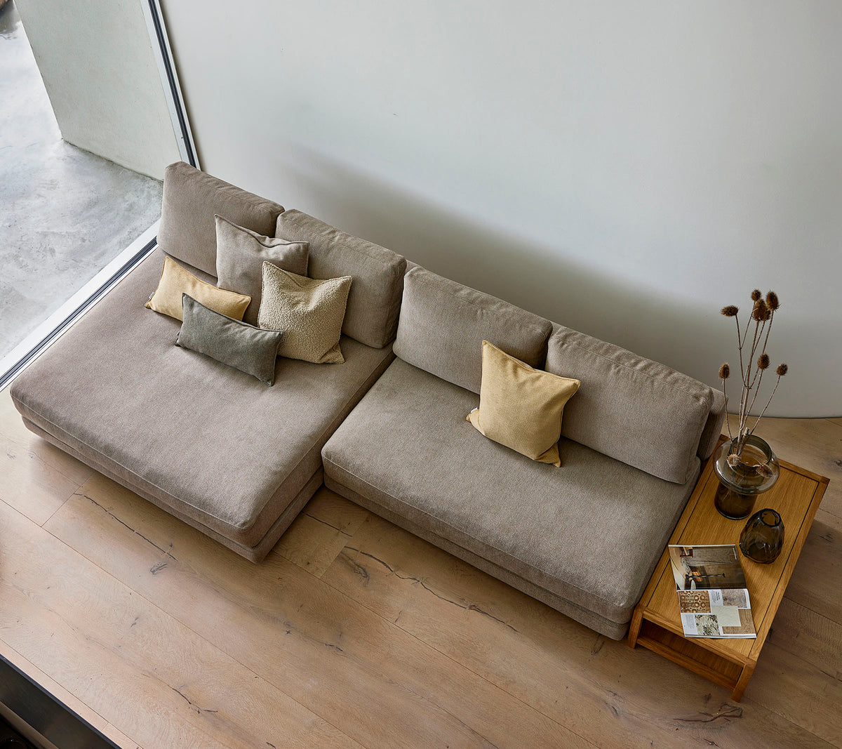 Un sofá elegante en un tono neutro, adornado con varios cojines decorativos, acompañado de una mesa auxiliar de madera con libros y un jarrón.