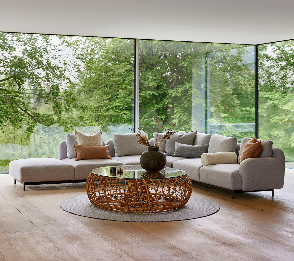 Área de salón moderna con un sofá modular con almohadas decorativas, una mesa de café redonda de mimbre y grandes ventanas de vidrio que muestran la vegetación.