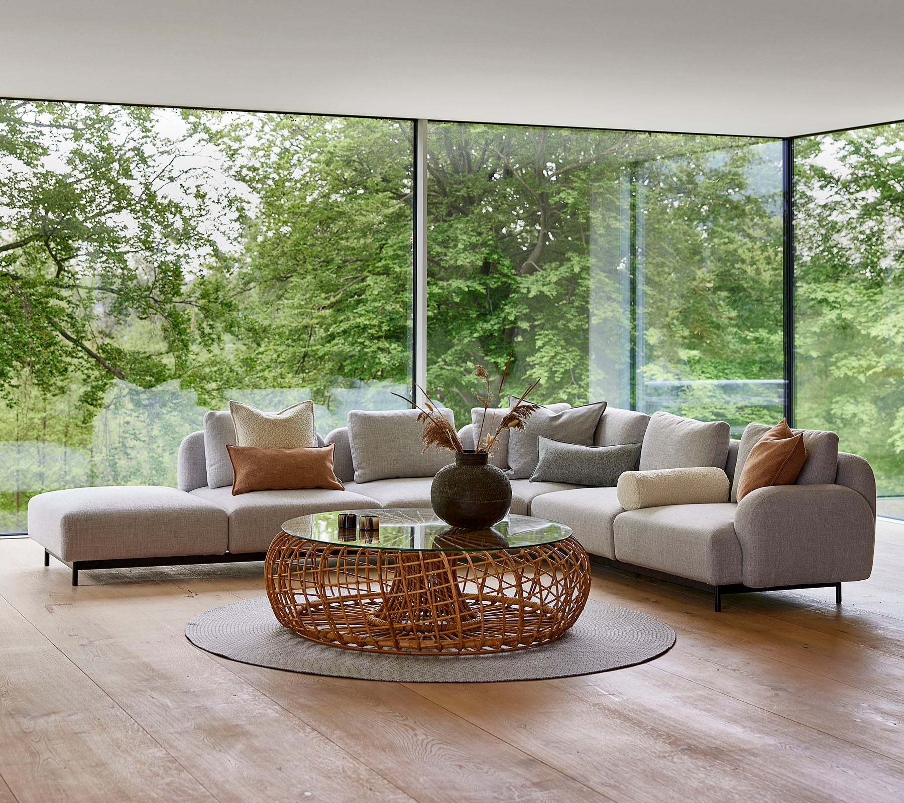 Área de salón moderna con un sofá modular con almohadas decorativas, una mesa de café redonda de mimbre y grandes ventanas de vidrio que muestran la vegetación.