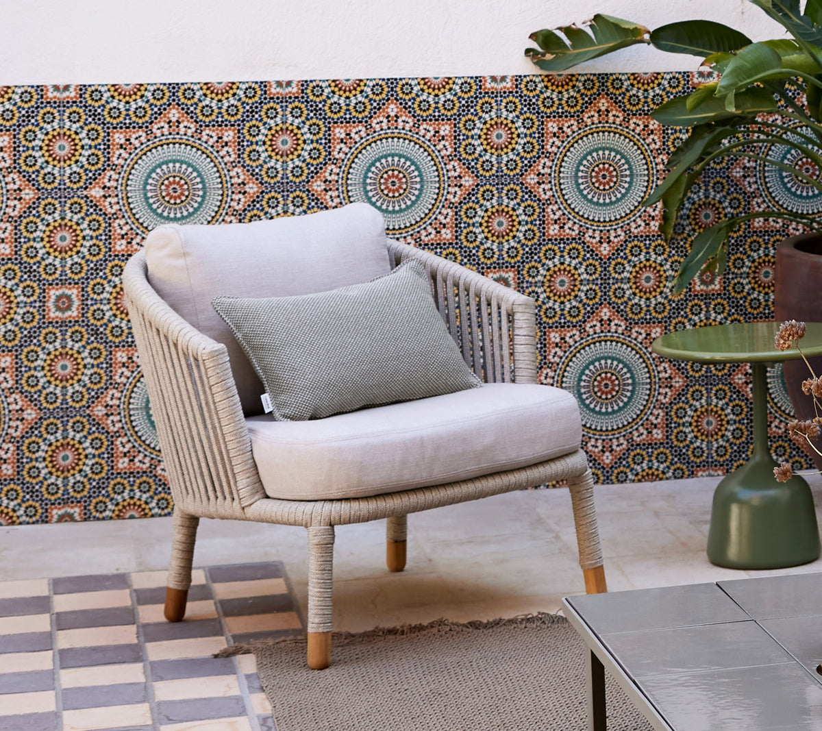 Una silla lounge elegante con un cojín suave y un cojín decorativo, ubicada contra un fondo estampado y rodeada de una planta en maceta.