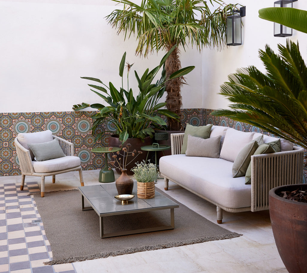 Zona de salón acogedora con un sofá claro, plantas en macetas, mesa decorativa y suelo texturizado, creando una atmósfera serena.