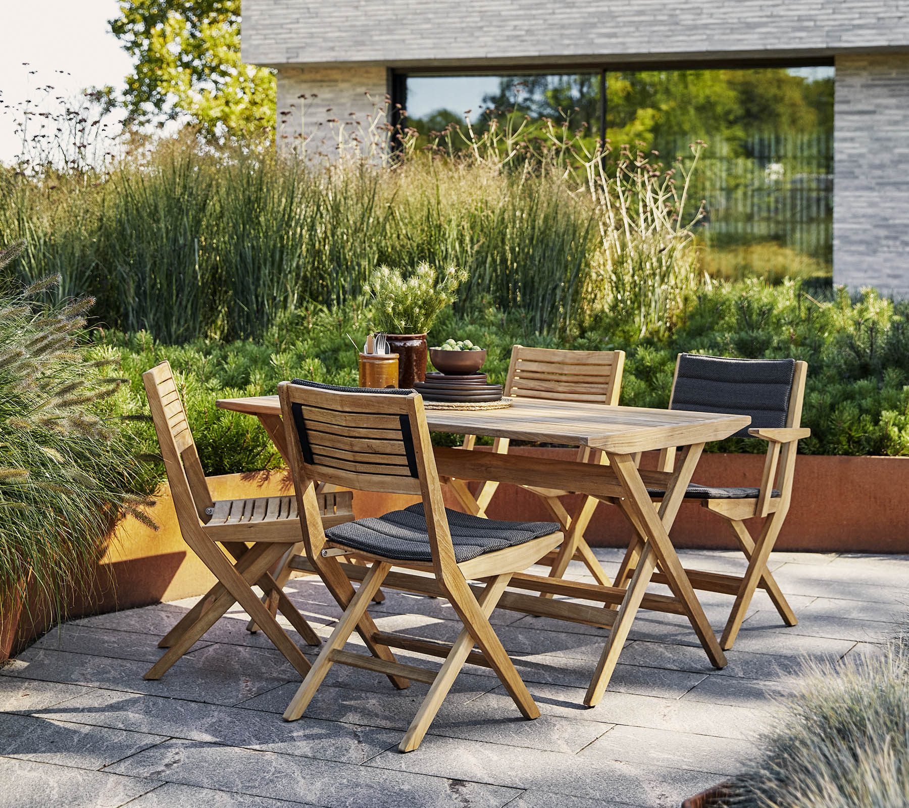 Mesa de madera elegante con sillas a juego rodeadas de exuberante vegetación y un telón de fondo arquitectónico moderno. Perfecta para reuniones al aire libre.
