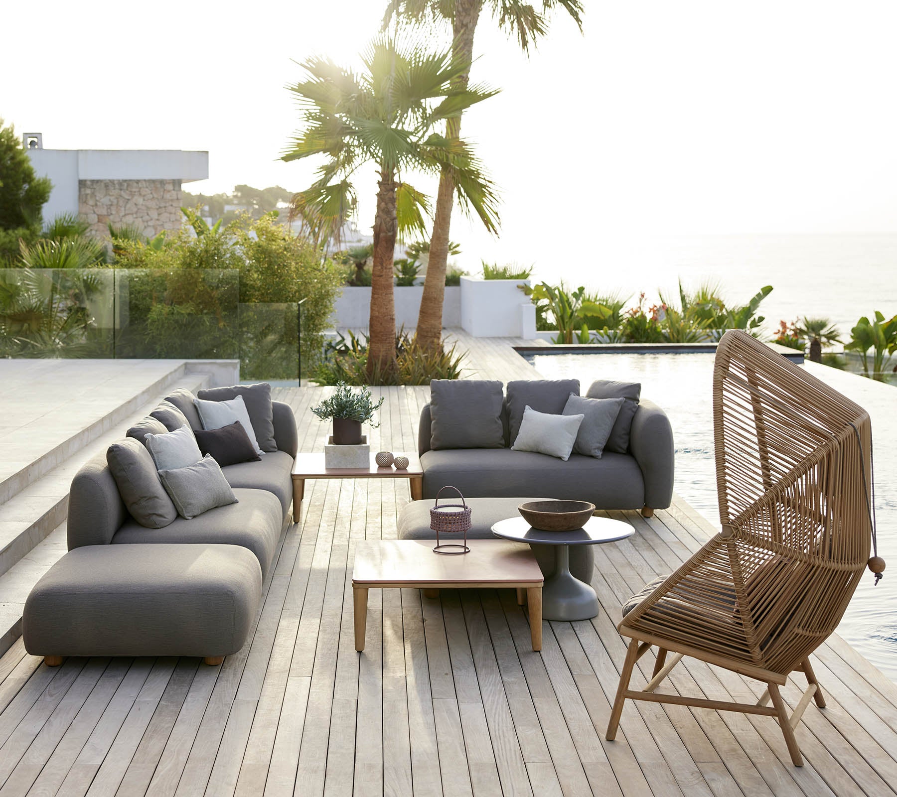 Zona de estar elegante con sofás grises y una silla de mimbre, complementada con una mesa de madera y vegetación exuberante de fondo.