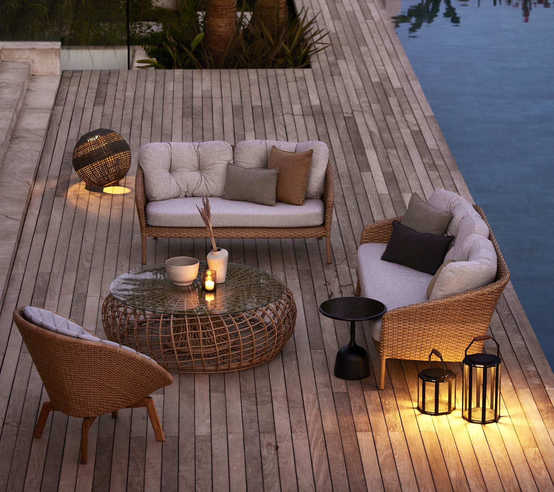 Zona de estar al aire libre acogedora con muebles trenzados, cojines suaves y iluminación ambiental al lado de una piscina en una cubierta de madera.
