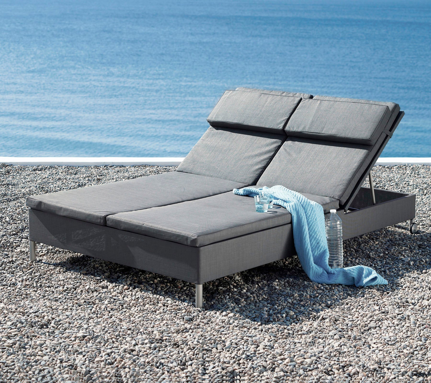 Una elegante silla de salón gris con respaldo ajustable, acompañada de una suave manta, junto a una tranquila vista al agua.
