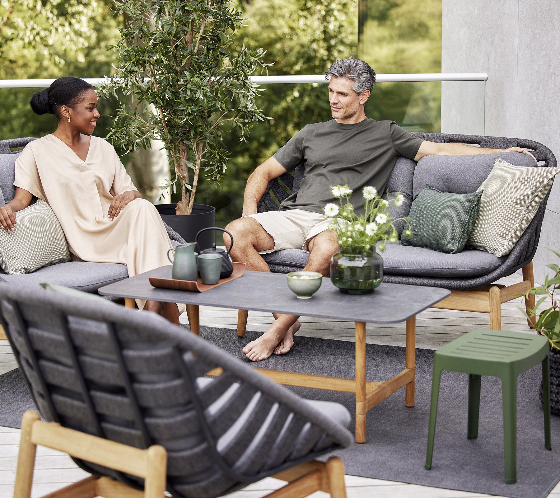Dos personas se relajan en un sofá exterior elegante, rodeados de vegetación y plantas decorativas, disfrutando de un momento pacífico juntos.